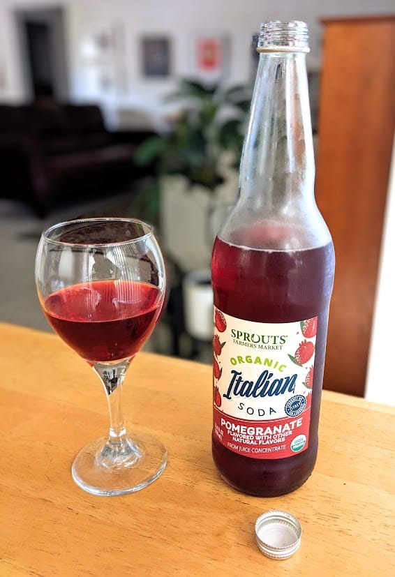 A bottle of Sprouts Pomegranate flavor Organic Italian Soda next to a wine glass full of red drink.