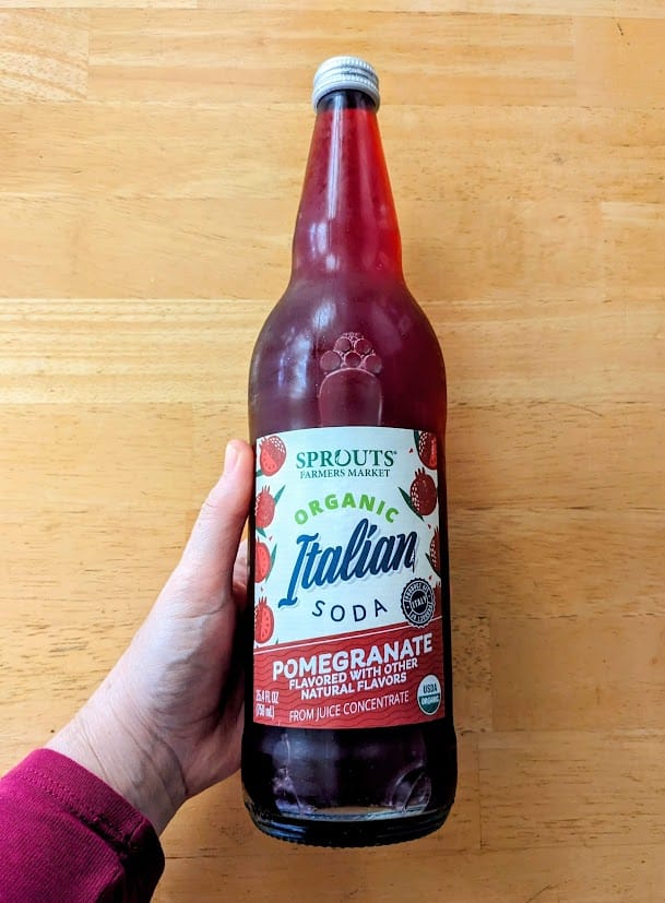 A hand holding a glass bottle of Sprouts Organic Pomegranate Italian Soda over a kitchen table.