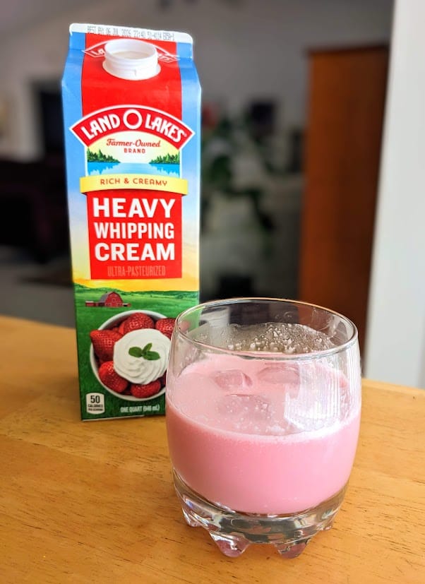 A quart of heavy whipping cream on a kitchen table next to a glass of pink cream soda.