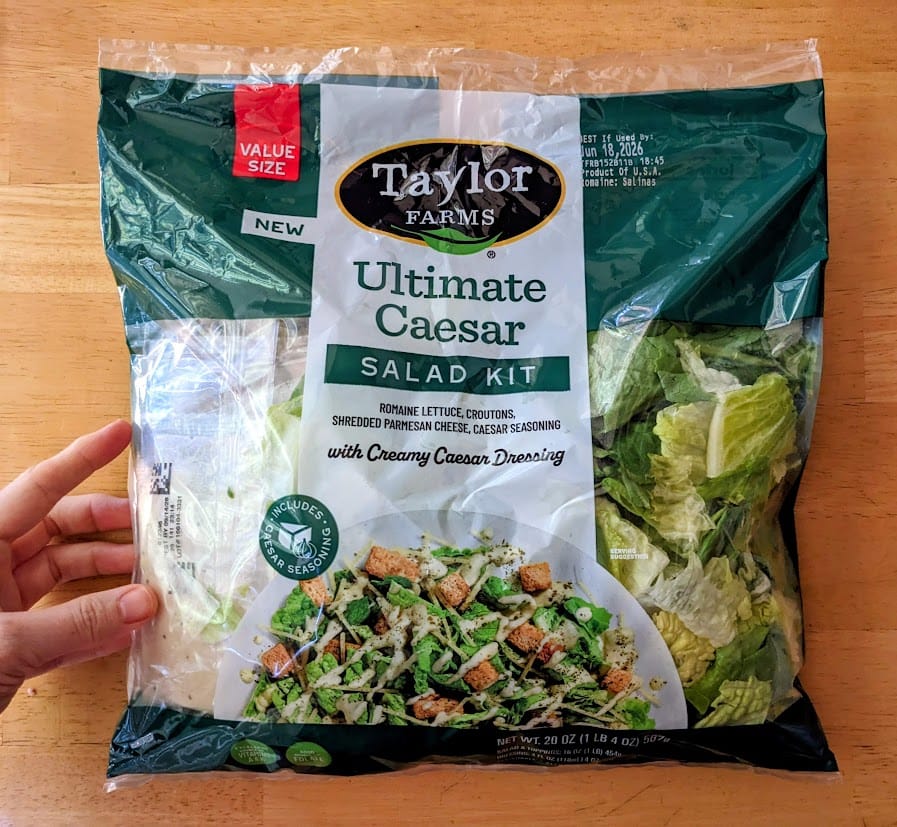 A hand holding a bag of Taylor Farms Ultimate Caesar Salad Kit from Costco.