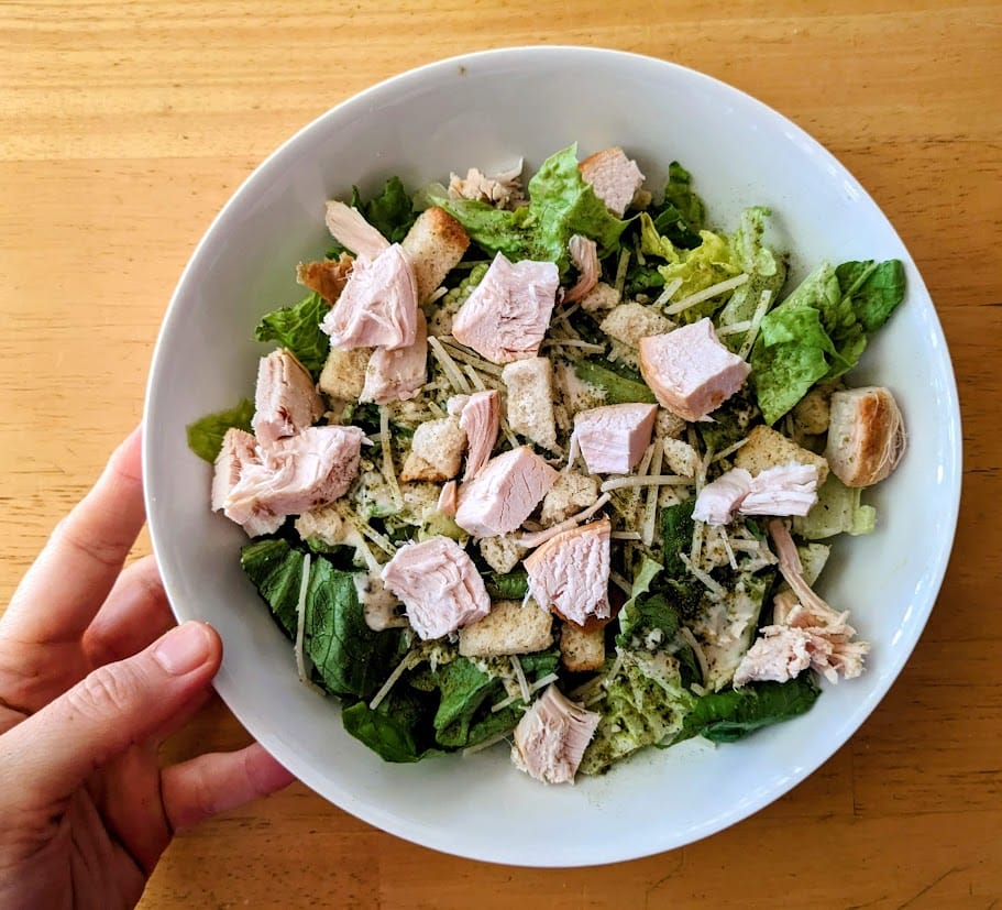 A hand holding a white bowl with caesar salad topped with cubes of Costco rotisserie chicken breast.
