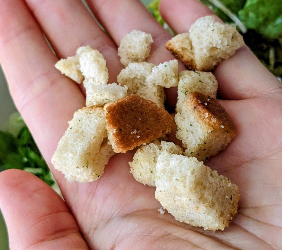 A hand holding croutons from a Taylor Farms Ultimate Caesar Salad Kit.