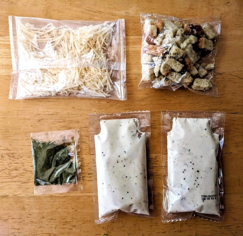 Plastic pouches of parmesan cheese, croutons, caesar dressing, and seasoning mix from a Taylor Farms salad kit.