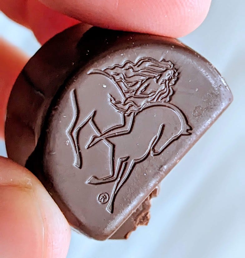 Fingers holding half a Godiva Masterpiece Ganache Chocolate, showing the embossed design on the top of a woman riding a horse.