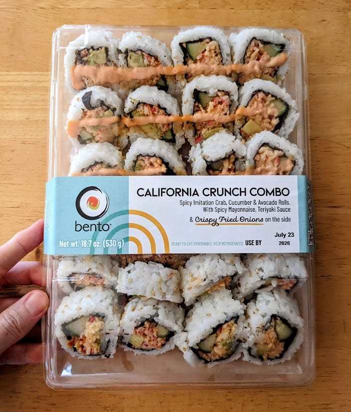 A hand holding a box of Costco Bento California Crunch Combo Sushi rolls.