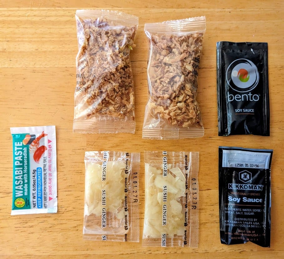 Condiments from Costco sushi, including wasabi paste, pickled ginger, soy sauce, and crunchy fried onions.