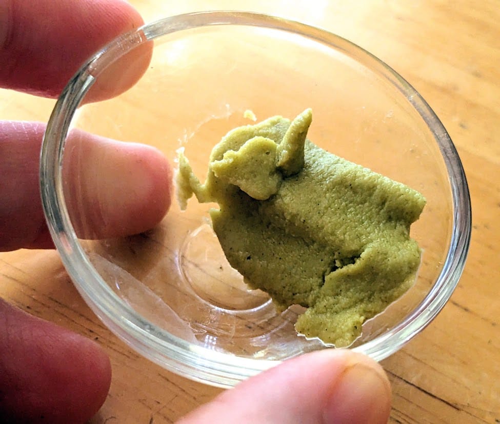 Fingers holding a small glass bowl with Costco sushi wasabi paste.
