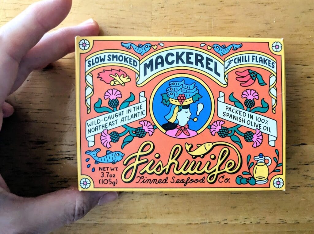 A hand holding a box of Fishwife Smoked Mackerel with Chili Flakes tinned seafood.