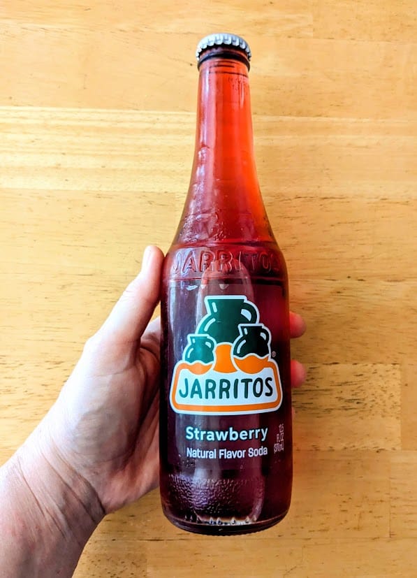 A hand holding a glass bottle of Jarritos Strawberry Mexican Soda.