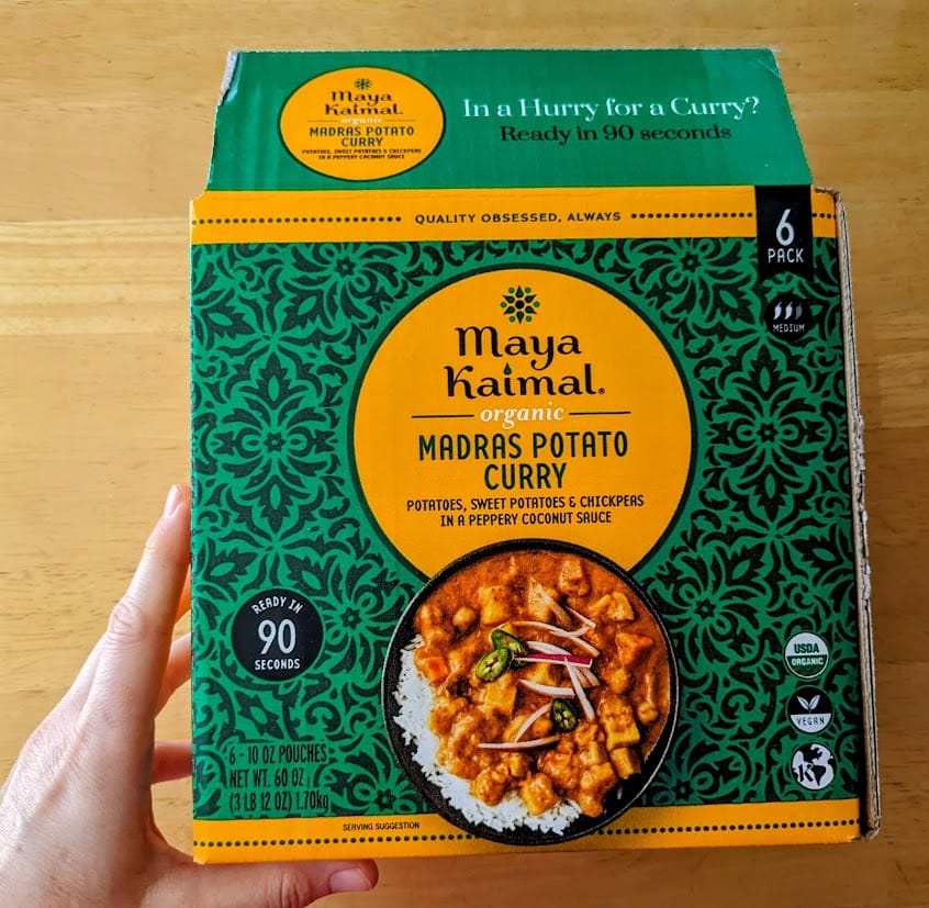 A hand holding a large box of Maya Kaimal Organic Madras Potato Curry from Costco.