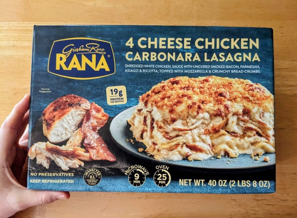 A hand holding a box of Rana 4 Cheese Chicken Carbonara Lasagna from Costco.