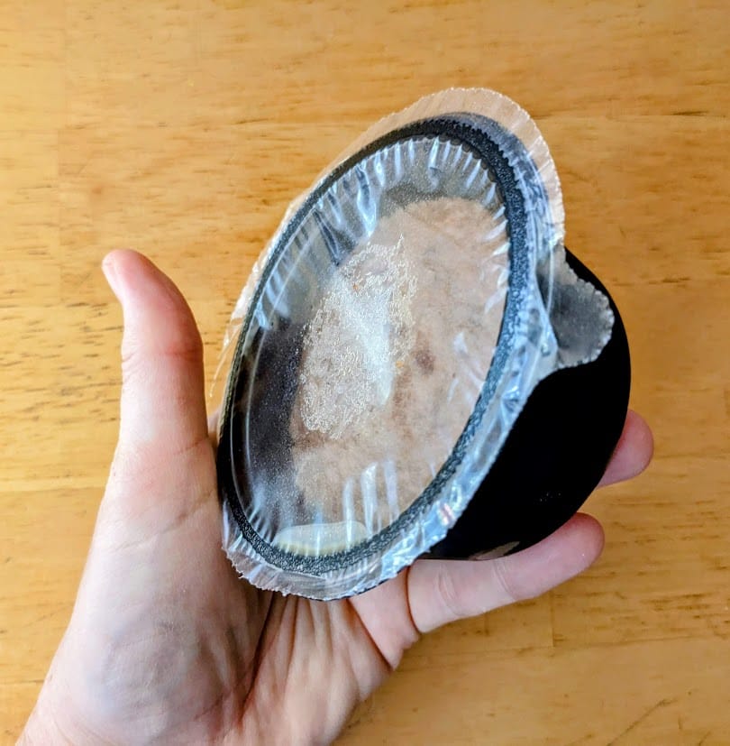 A hand holding a black plastic sealed cup with The Sticky Toffee Pudding Company British cake from Costco.