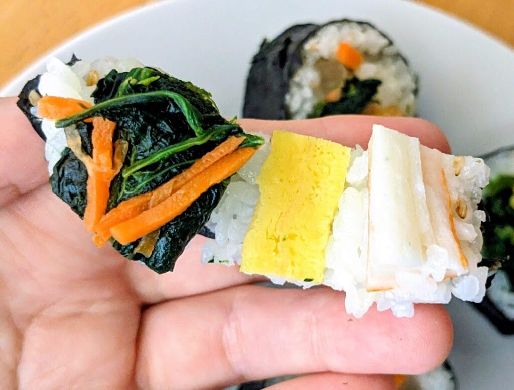 A hand holding a peeled open piece of Aldi Kimbap, showing the spinach, egg, and krab.