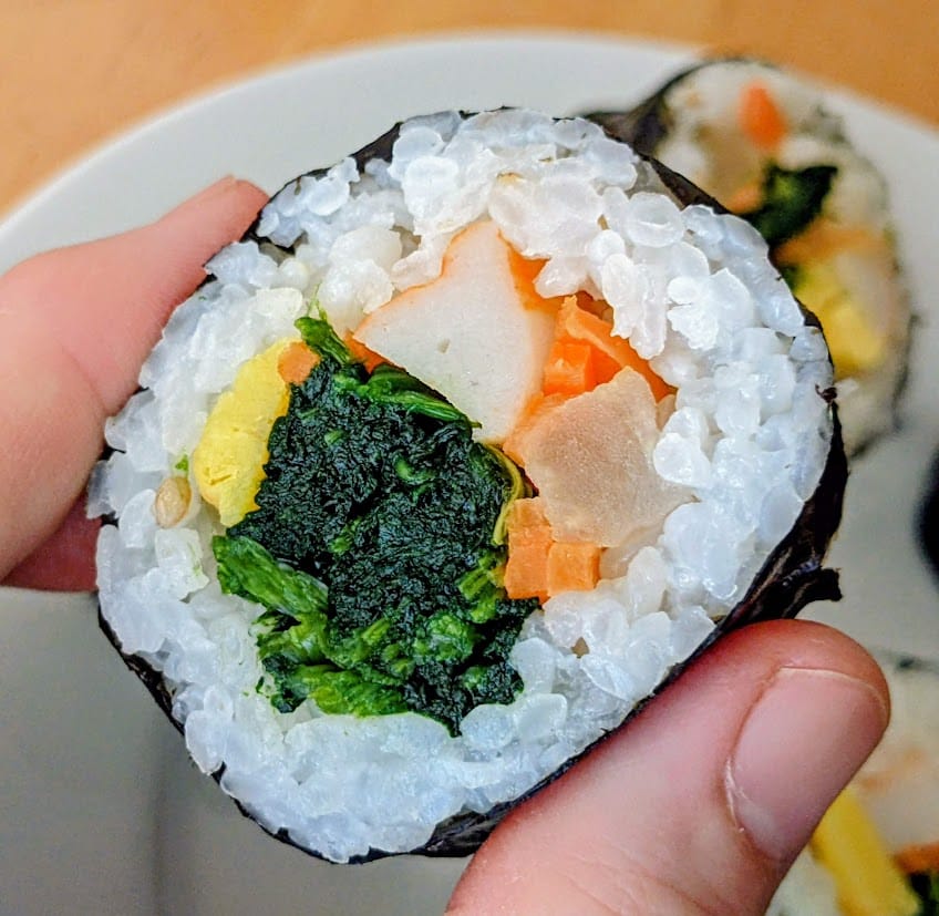 Fingers holding a piece of Aldi Kimbap, Traditional flavor, showing the spinach, radish, carrots, and egg.