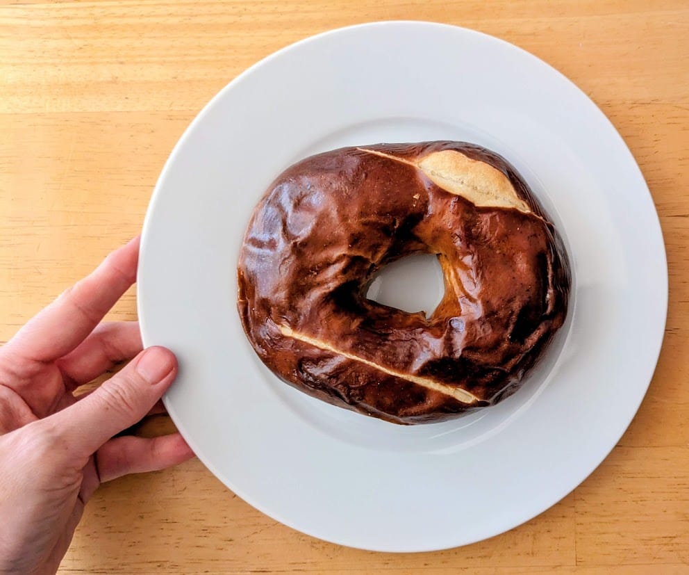 A hand holding a white plate with a whole Costco Pretzel Bagel.