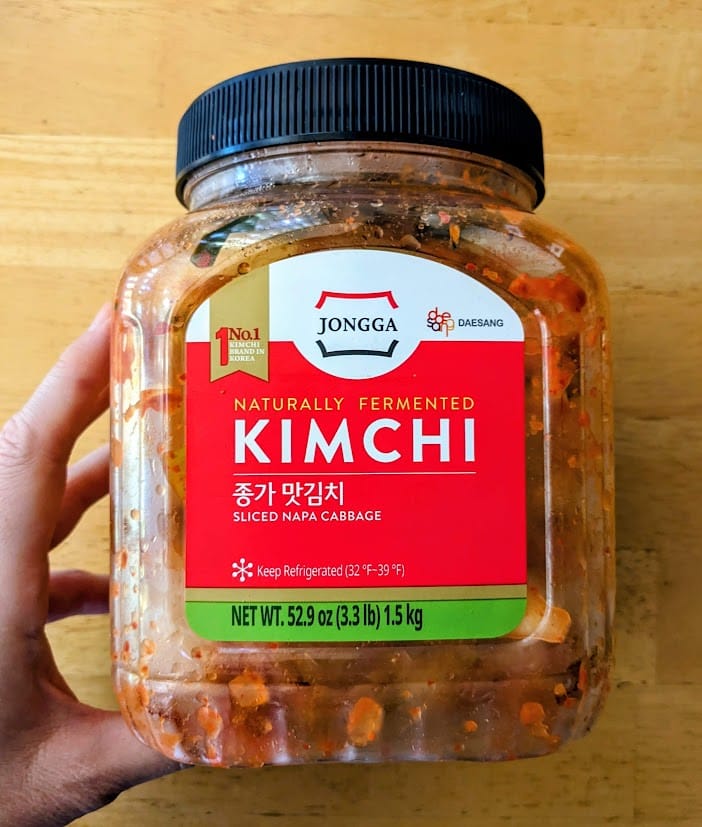 A hand holding a plastic jar of Jongga Naturally Fermented Korean Kimchi from Costco.