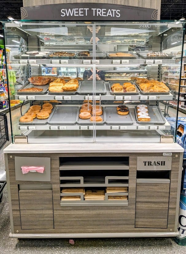 A glass "Sweet Treats" case inside a Publix Bakery, with self-serve bags, tissue paper, and trash receptacle.