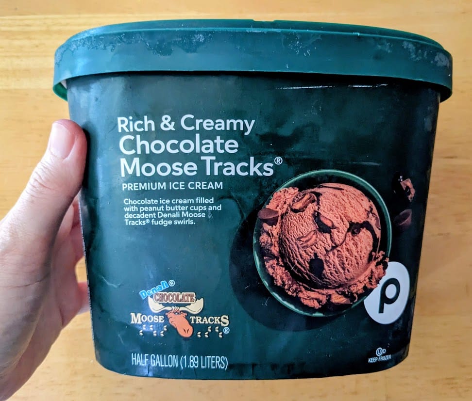 A hand holding a dark green half gallon carton of Publix Chocolate Moose Tracks Ice Cream.