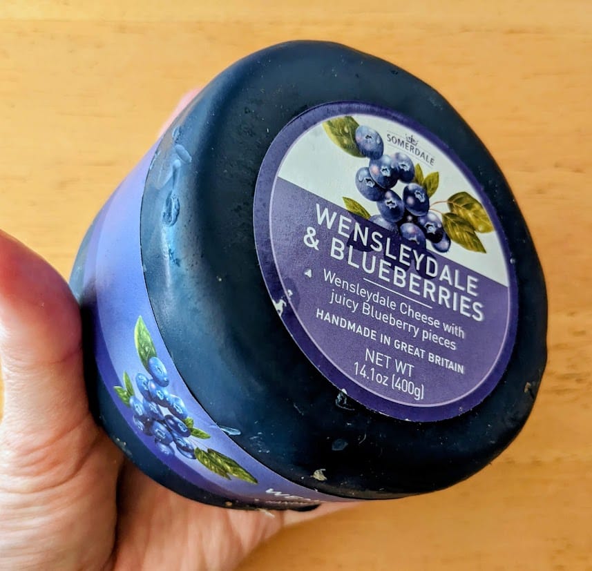 A hand holding a blue wax-wrapped wheel of Somerdale Wensleydale & Blueberries Cheese from Costco.