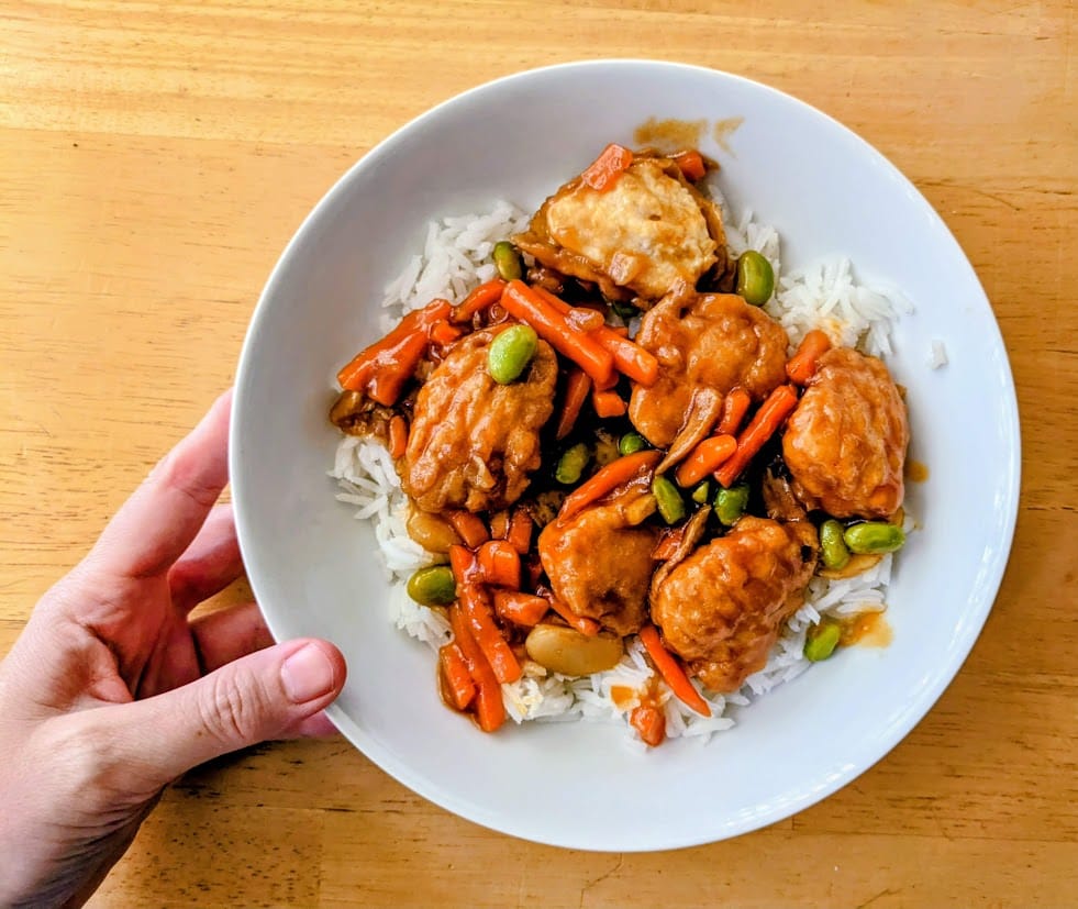 A hand holding a white bowl of basmati rice topped with PF Chang's Orange Chicken frozen meal.