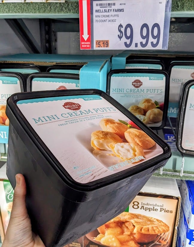 A hand holding a large frozen tub of Mini Cream Puffs inside a BJ's Wholesale Club warehouse.