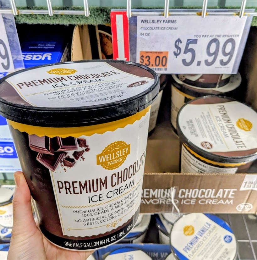 A hand holding a half gallon of Wellsley Farms Premium Chocolate Ice Cream inside a BJ's Wholesale Club.