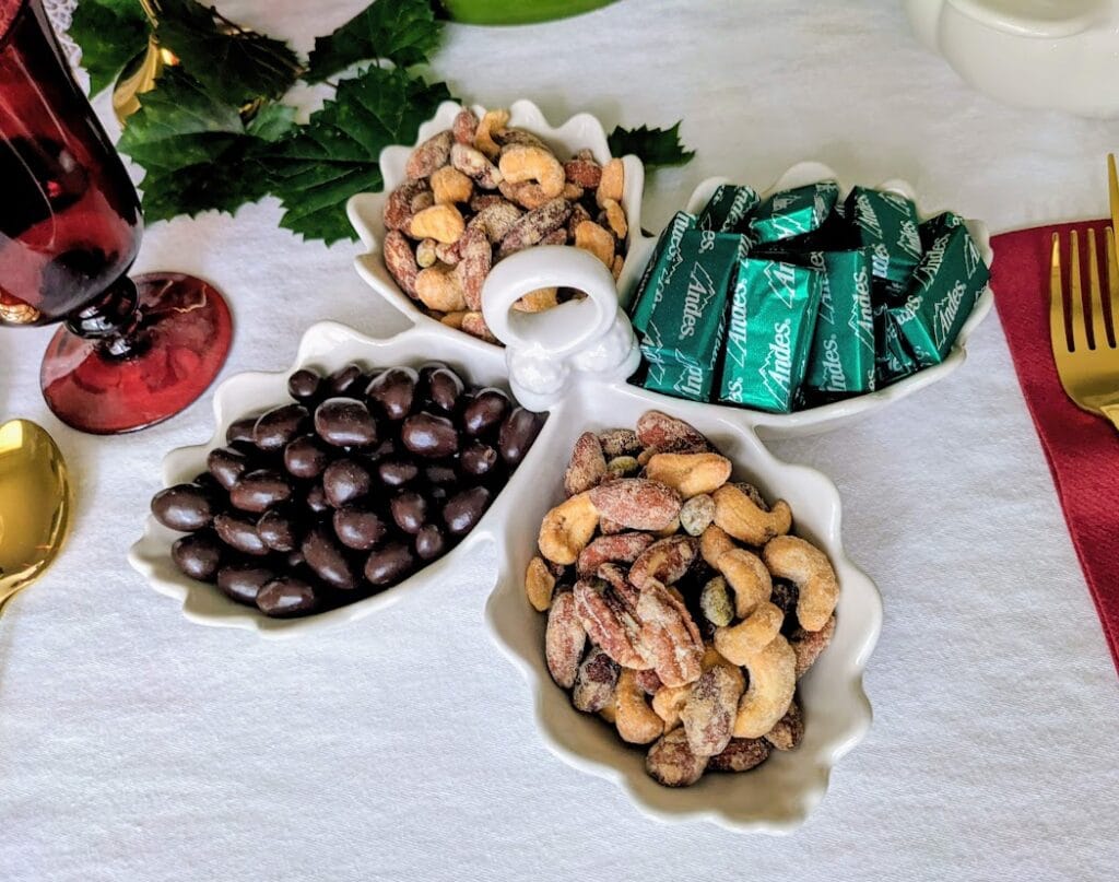 A ceramic acorn and leaf-shaped four compartment server filled with nuts, chocolate, and mints, sitting on a holiday dinner table.
