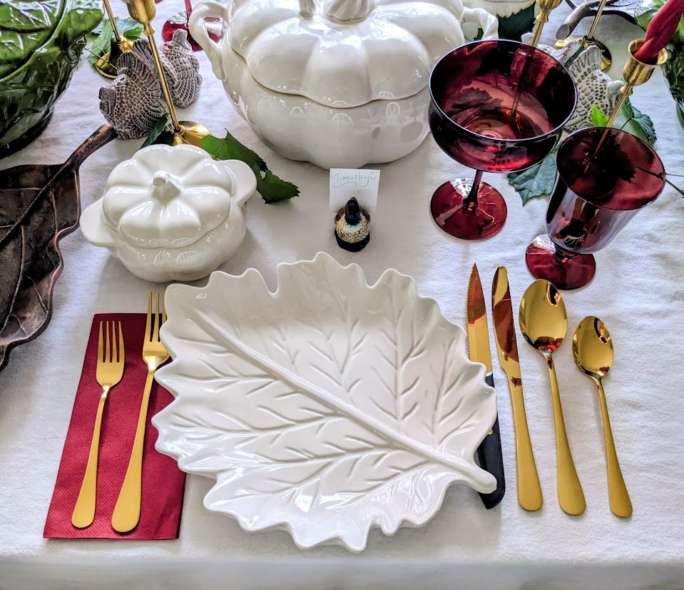 A holiday table place setting with gold flatware, dark red glasses, ivory pumpkins, and a leaf shaped plate.